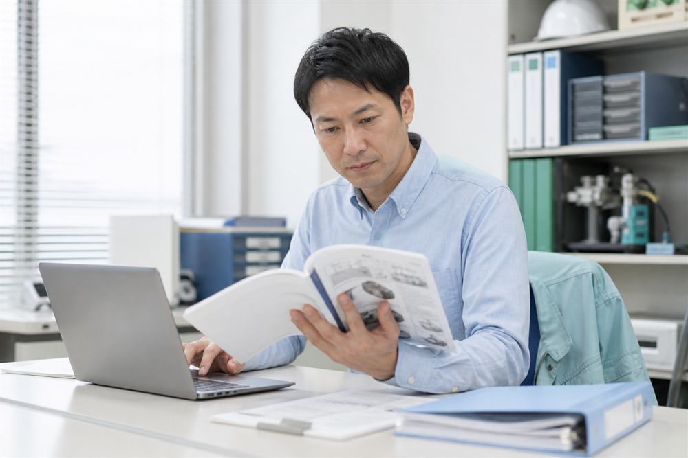 ノートPCとマニュアルを見比べながら資料を確認する技術文書担当者